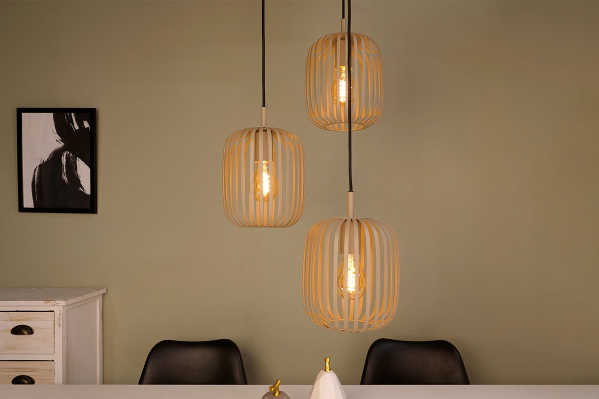 Drie hangende pendellampen met een kap van metalen staven in een moderne eetkamer boven een eettafel met zwarte stoelen.