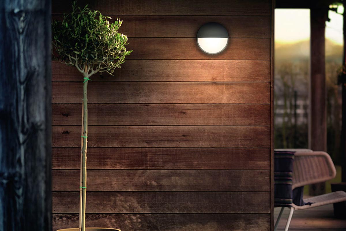 Een halfronde wandlamp aan een houten wand naast een kleine boom.