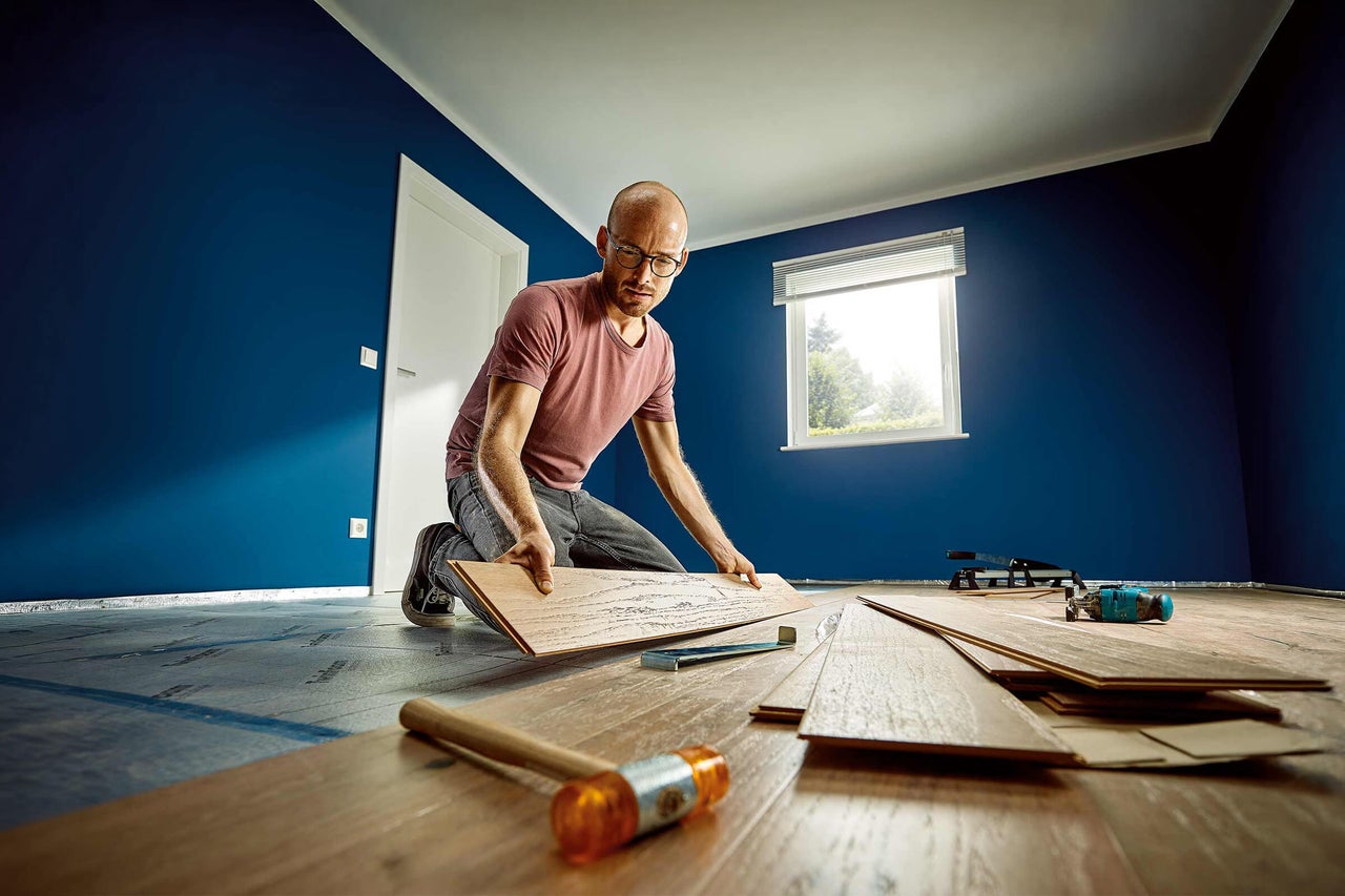 Een man legt een lichtbruine laminaatvloer in een lichte kamer met blauwe muren. Leggereedschap ligt op de voorgrond klaar.