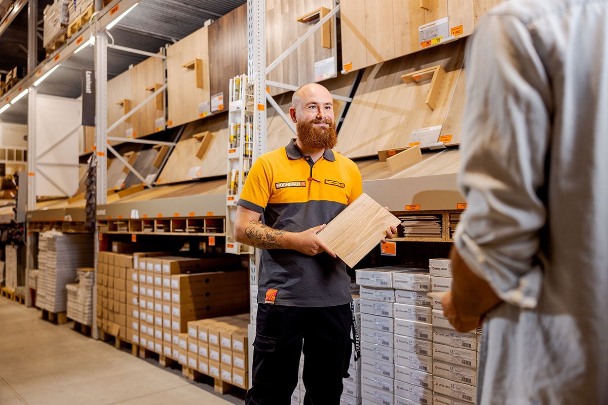 Een Hornbach medewerker houdt een laminaatmonster vast, op de achtergrond een schap met laminaat.