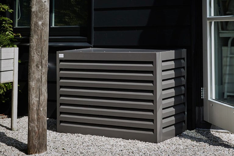 Metalen airco ombouw met horizontale lamellen voor een zwarte muur op grind.