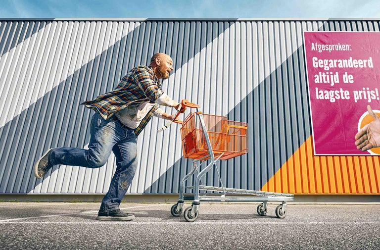Een man in geruit overhemd en spijkerbroek rent met een oranje winkelwagen voor een Hornbach-gevel met een bord voor de laagsteprijsgarantie.