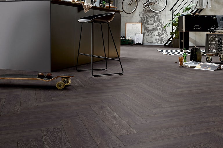 Interieur met donkere houten vloer in visgraatmotief, aangevuld met een kookeiland, een barkruk en decoratieve elementen