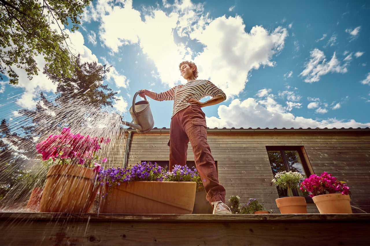 Een persoon bewatert bloemen in potten op een houten terras met een metalen gieter voor een houten huis.