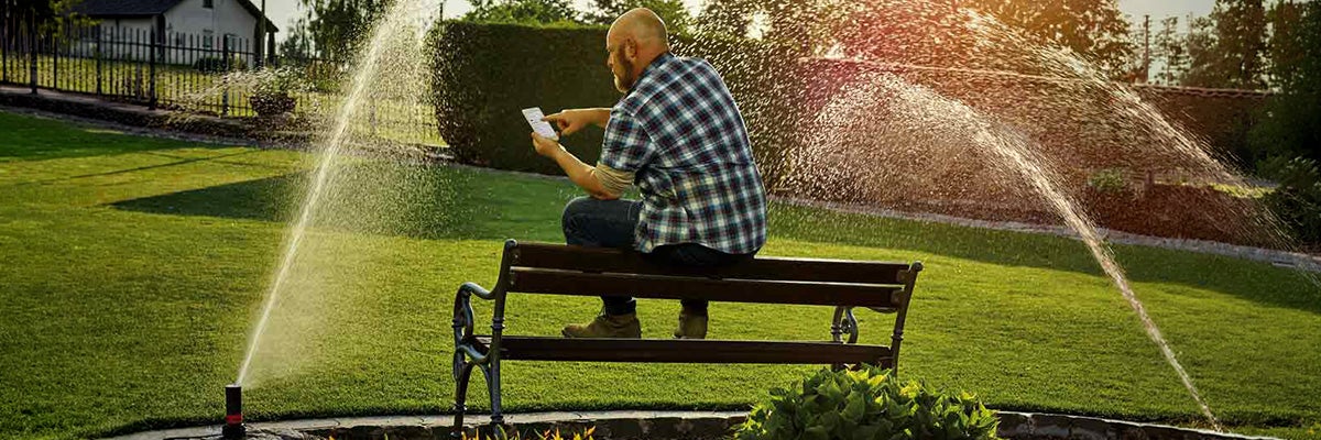 Een man zit op een tuinbank op een gazon en bedient het automatische besproeiingssysteem met verzonken sproeiers via zijn smartphone.