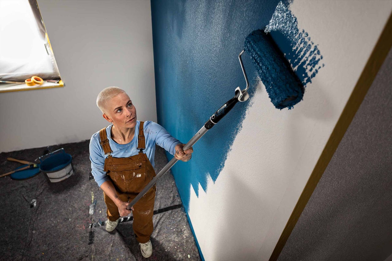 Een persoon met kort haar en een werkbroek schildert een muur met een verfroller en telescoopsteel.