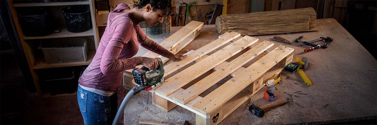 Een vrouw bewerkt een houten pallet met een accu-decoupeerzaag in een werkplaats met diverse gereedschappen.