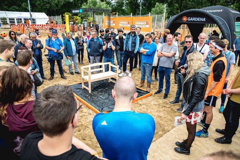 Een groep mensen staat buiten rond een houten bank, met Hornbach- en Gardena-reclame-elementen op de achtergrond.
