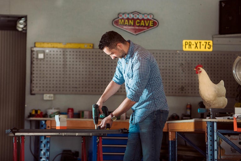 Man werkt met een accuschroevendraaier in een werkplaats. Op de achtergrond een gereedschapswand, een bord met de tekst Man Cave en een kip.