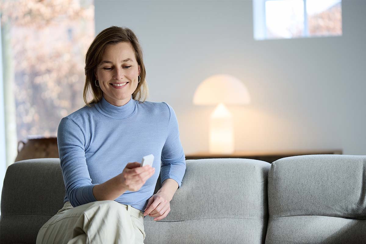 Een glimlachende vrouw zit op een bank en bedient een klein smart home besturingsapparaat in een modern ingerichte woonkamer.
