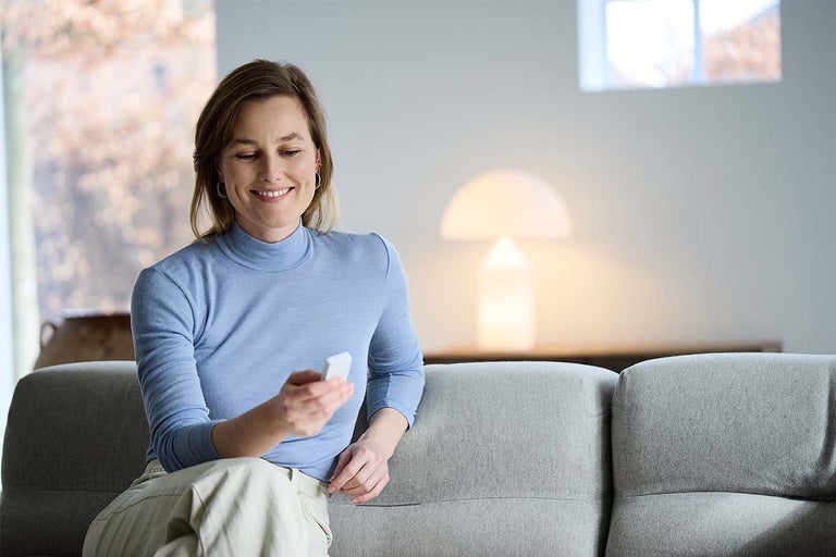 Een glimlachende vrouw zit op een bank en bedient een klein smart home besturingsapparaat in een modern ingerichte woonkamer.