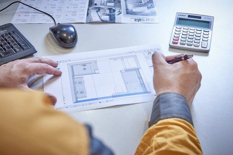 Een persoon bekijkt een badkamerplan op papier met computeraccessoires en een rekenmachine op een tafel.