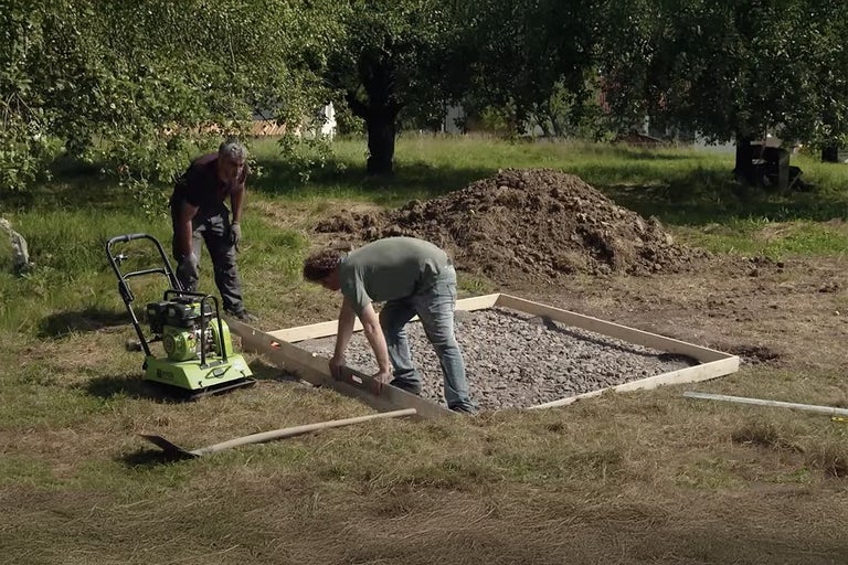 Tuinscene met personen die een fundering voorbereiden met een trilplaat en een schop.