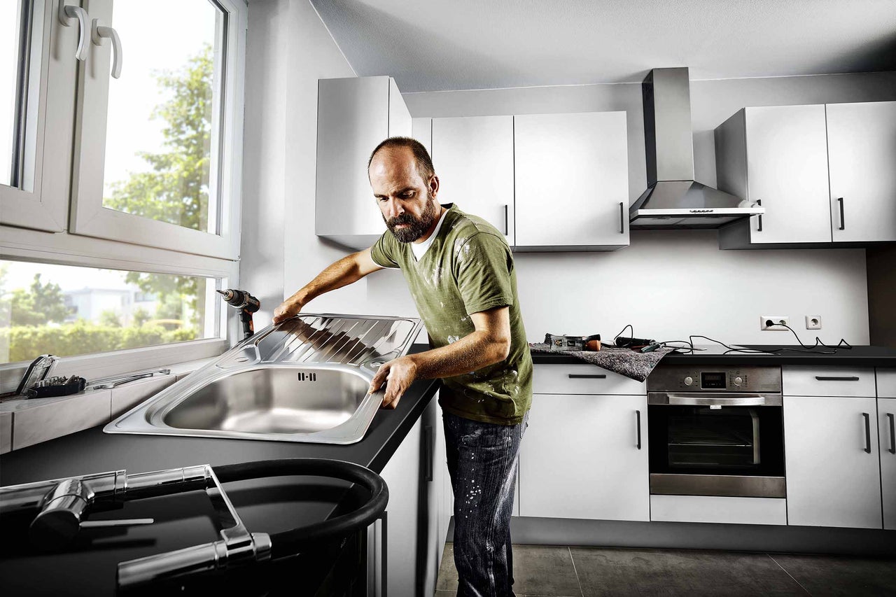 Klusjesman installeert spoelbak in moderne keuken met witte kasten en donker aanrechtblad.