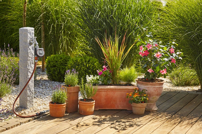 Tuin met waterkraan, besproeiingscomputer, tuinslang en beplante terracotta potten op een houten terras