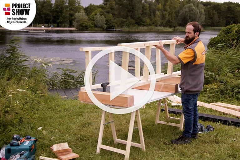 Een man werkt aan een houtproject aan het meer. Op de voorgrond staat een werkbank met een houten frame. Op de achtergrond is een meer te zien.