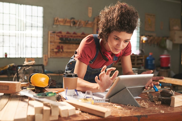 Vrouw met tablet in een werkplaats, omgeven door gereedschap en materialen voor houtbewerking.