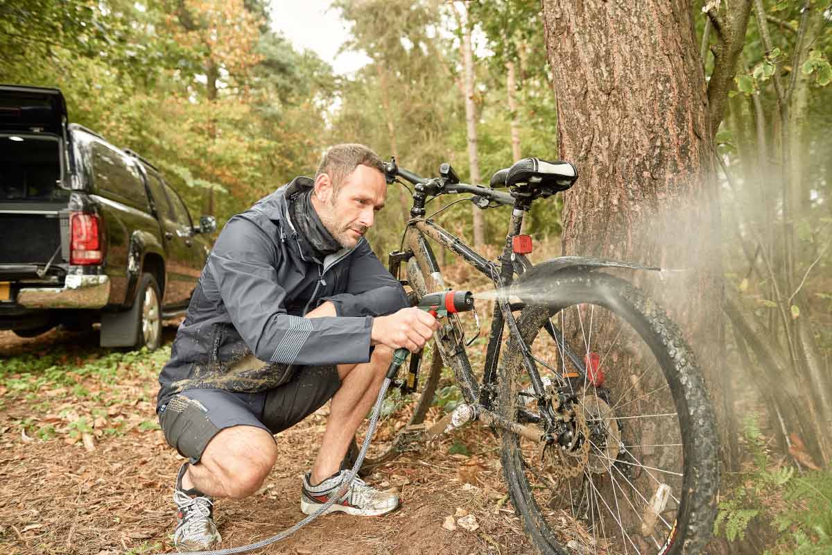 Man reinigt een vuile fiets met een tuinslang in het bos.