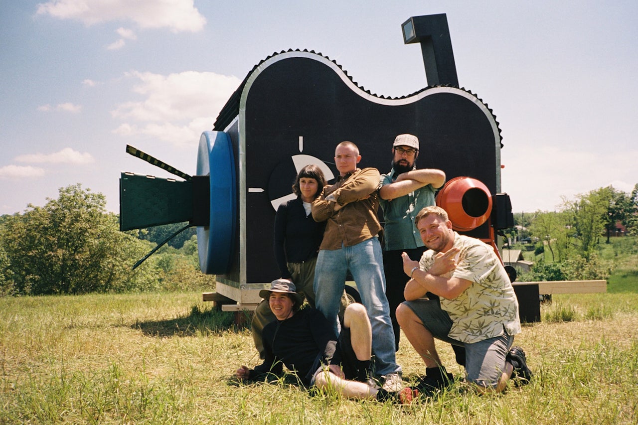 Een groep van vijf personen poseert voor een grote, artistieke constructie op een aanhanger in een groen veld.