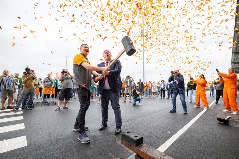 Twee personen slaan met een voorhamer op een ondergrond onder confetti, omringd door een menigte.