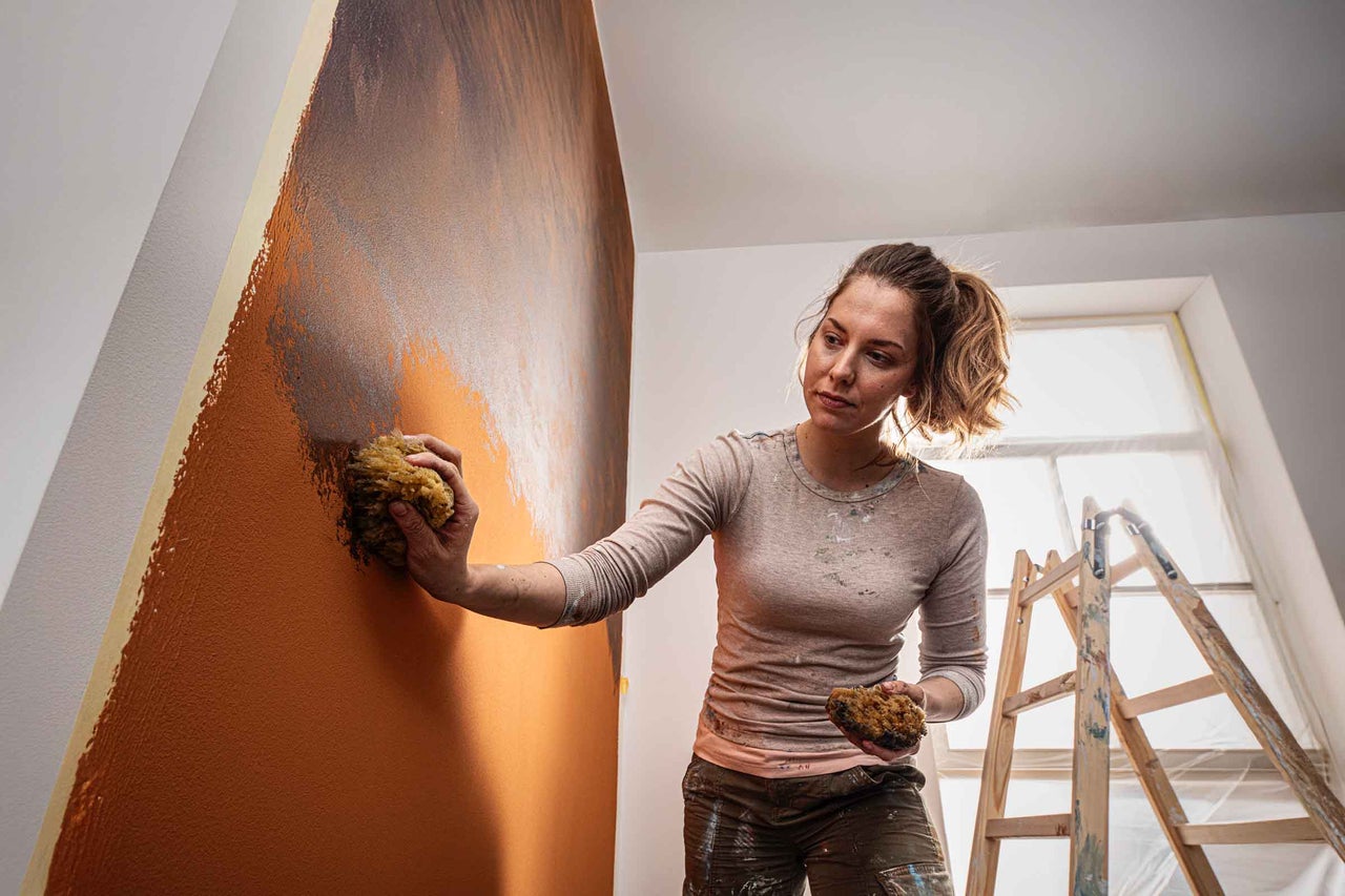 Een vrouw brengt met een natuurlijke spons muurverf aan op een oranje wand met de sponstechniek. Op de achtergrond staat een houten ladder.