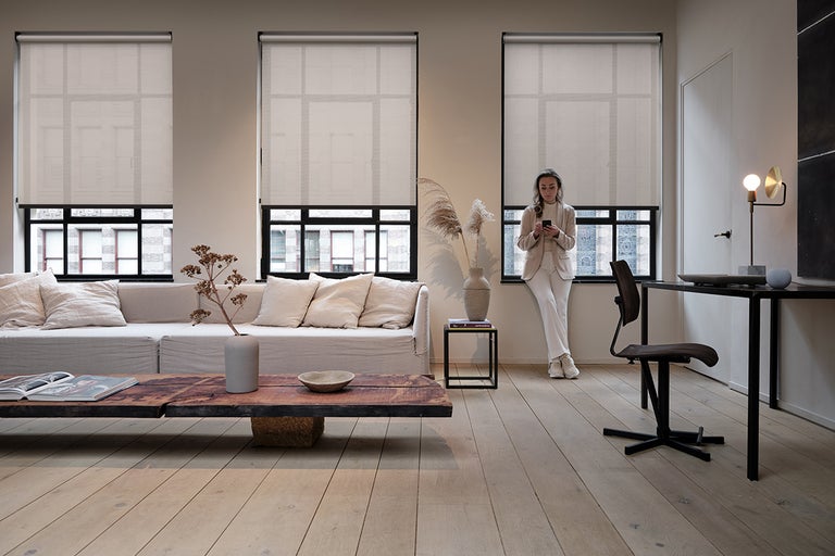 Woonkamer met drie ramen met grijze rolgordijnen, een bank en een bureau.