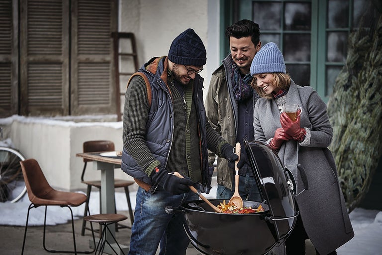 Drie mensen staan rond een kogelbarbecue waar voedsel op wordt bereid