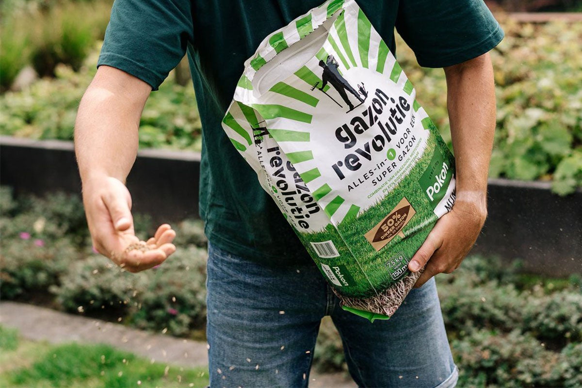 Een persoon houdt een pak Pokon gazonzaden vast en strooit de zaden uit.