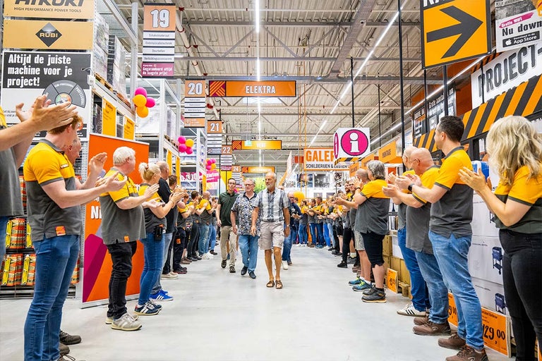 Interieur van een Hornbach bouwmarkt met werknemers die klanten toejuichen, samen met de afdelingsaanduidingen Elektra, Galerie en Montage