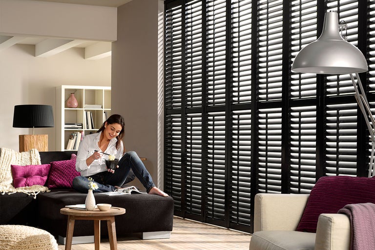 Woonkamer met zwarte luiken, bank, lamp en vrouw.