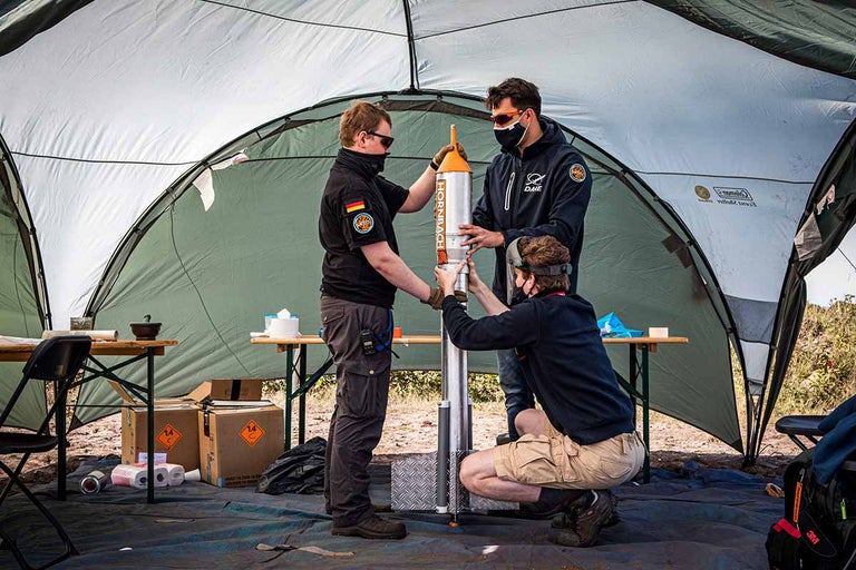 Drie mensen monteren een raket onder een tent. Tafels en dozen zijn zichtbaar.