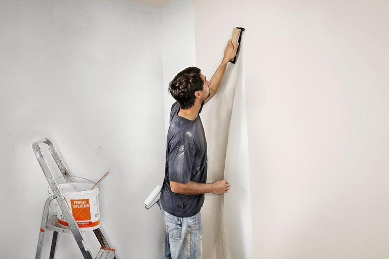Man behangt een muur met een behangborstel, op de achtergrond staan een ladder en een emmer behanglijm.