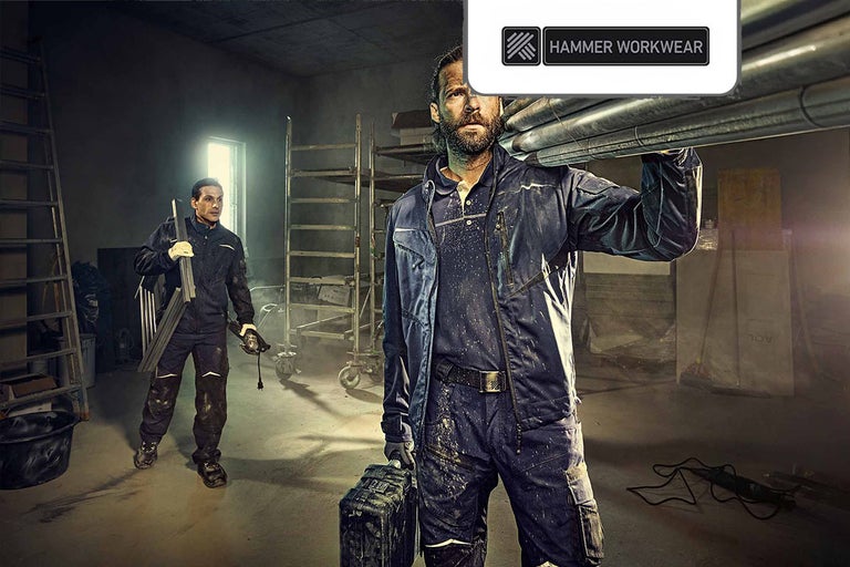 Twee vakmensen dragen werkkleding van het merk Hammer Workwear in een werkplaats. De voorste man draagt pijpen op zijn schouder en een gereedschapskist in zijn hand, terwijl de achterste man een metalen frame en een elektrisch gereedschap draagt. Op de achtergrond bevinden zich rekken en een ladder.