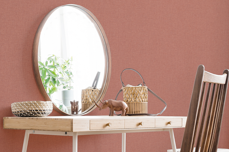 Gestructureerd vliesbehang in oudroze op een muur achter een houten bureau met een ronde spiegel en decoratieve voorwerpen.
