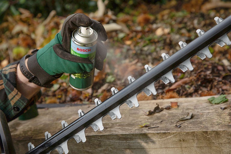 Een persoon spuit Bosch onderhoudsspray op het mes van een heggenschaar.