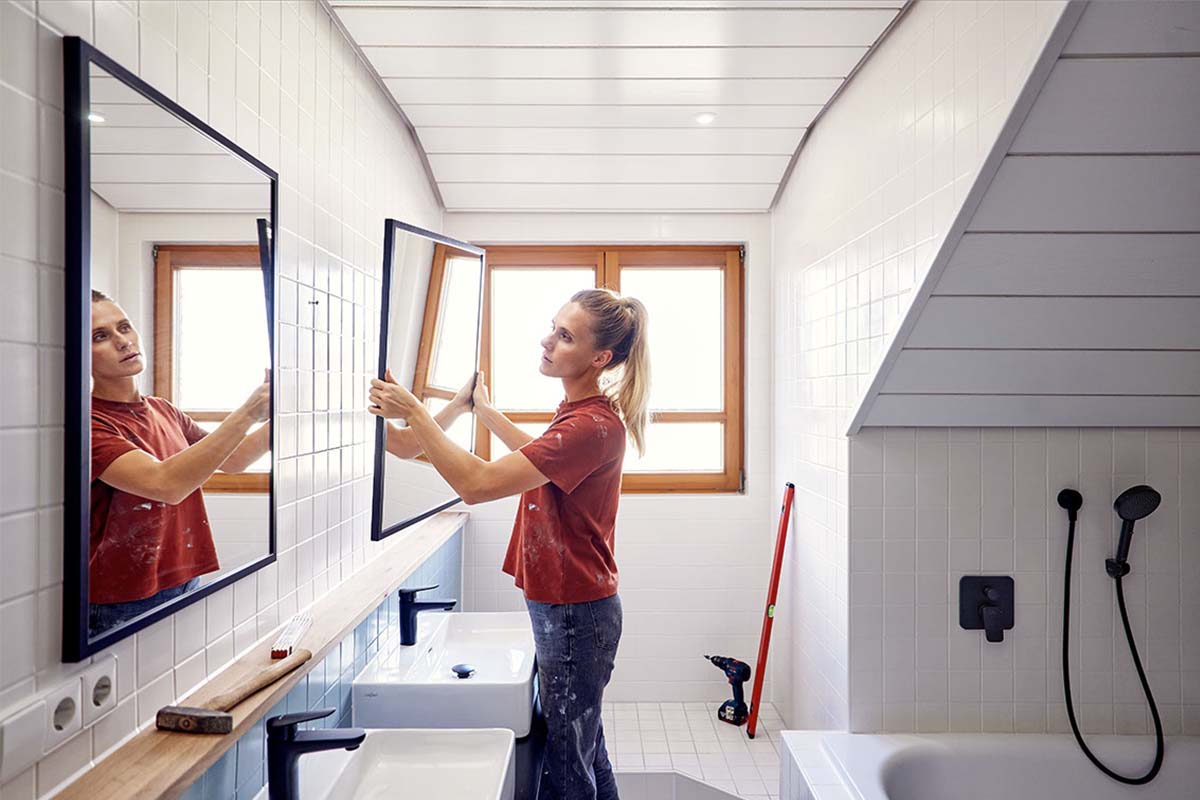 Een vrouw monteert een wandspiegel met zwart frame in een modern gerenoveerde badkamer met witte tegels.