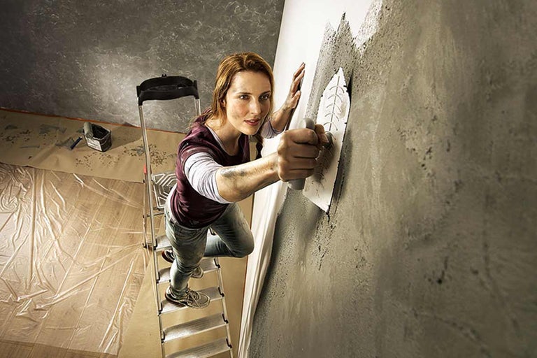 Vrouw op een ladder die met een troffel muurvuller op een muur aanbrengt tijdens het renoveren van een kamer.