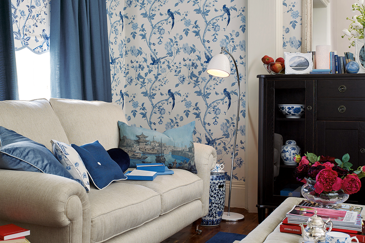 Woonkamer met vliesbehang met bloemen- en vogelpatroon, bijpassende gordijnen, beklede bank en dressoir.