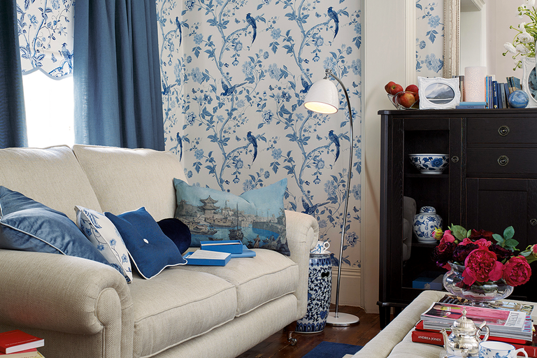 Woonkamer met vliesbehang met bloemen- en vogelpatroon, bijpassende gordijnen, beklede bank en dressoir.