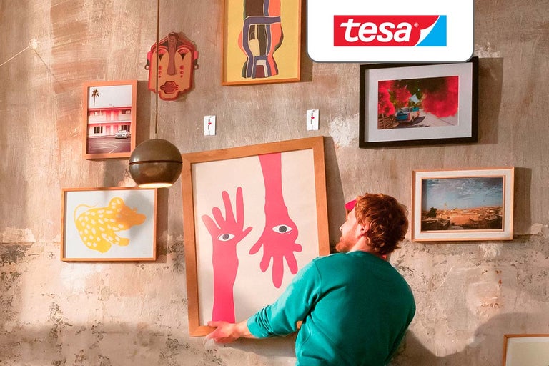Een man bevestigt fotolijsten aan een muur met structuur met behulp van klevende spijkers. tesa Logo.
