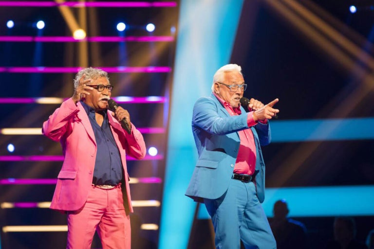 Twee mannen in pak zingen op een podium met microfoons.