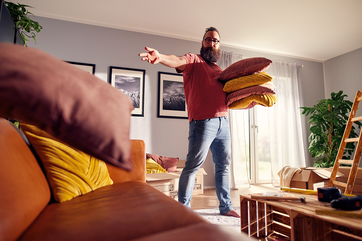 Een man draagt kussens in een lichte, ruime woonkamer tijdens een verhuizing.