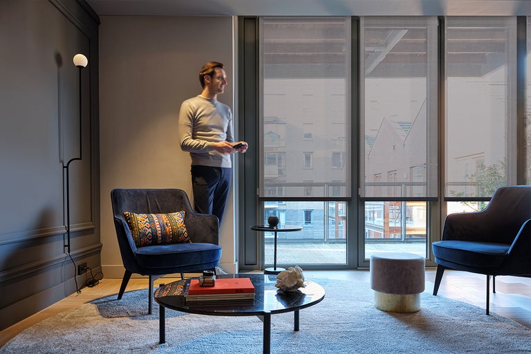 Woonkamer met fauteuil, salontafel, vloerlamp en rolgordijnen voor de ramen.