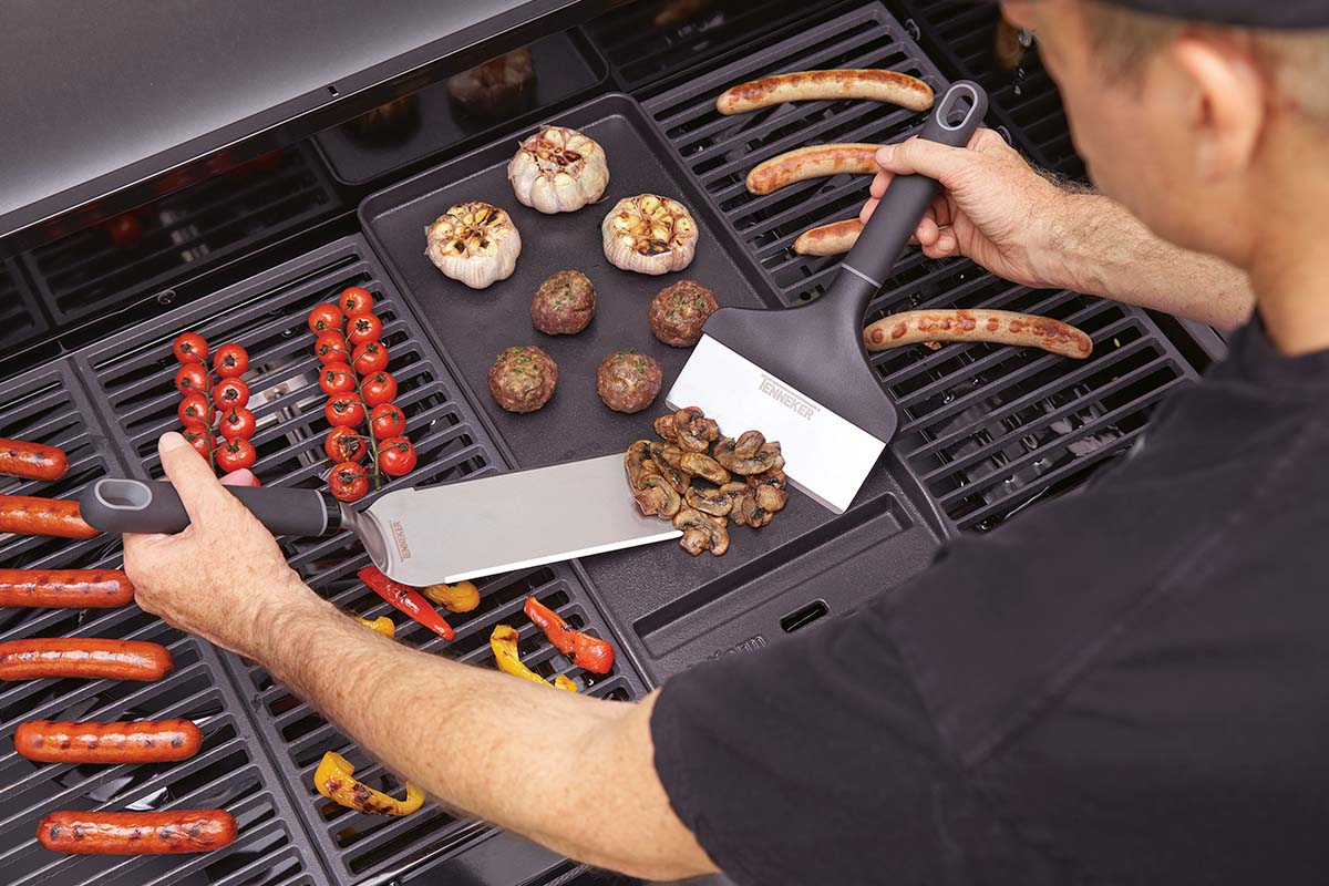 Op een grill liggen worstjes, tomaten, paprika's, champignons, knoflook en gehaktballetjes, die worden verplaatst met twee barbecuespatels.
