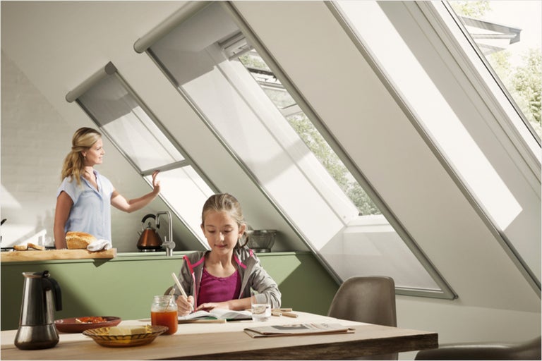 Dakramen met rolluik in een lichte ruimte, terwijl een vrouw het rolluik bedient en een meisje aan tafel zit