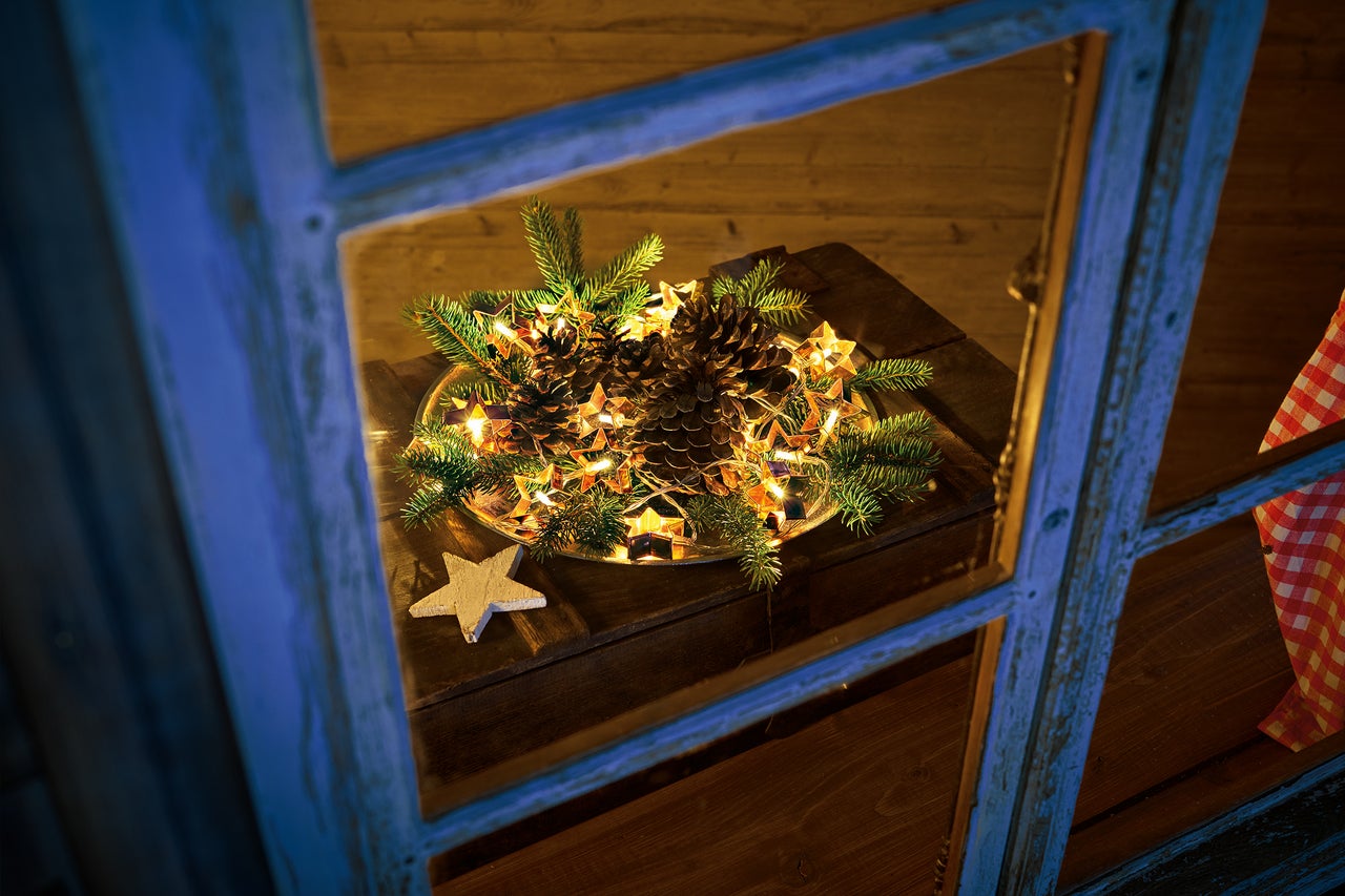 Decoratie met dennenappels, dennennaalden en sterrenlichtjes op een bord, gezien door een raam.