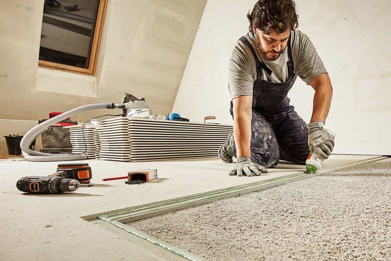 Een man dicht een droge dekvloer af met een afdichtmiddel. Gereedschap en materiaal liggen verspreid op de vloer.
