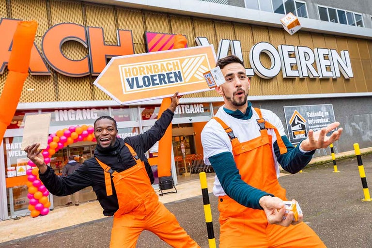 Twee medewerkers in tuinbroeken voor de Hornbach vestiging met een bord met de tekst vloeren.