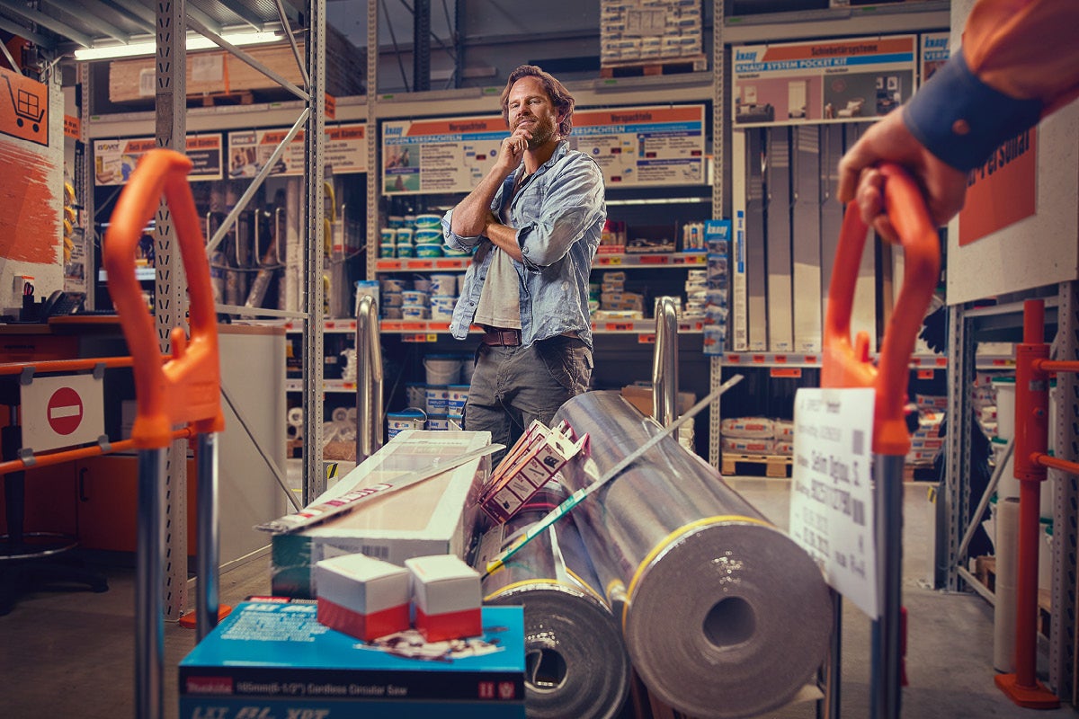 Een nadenkende man in een lichte, georganiseerde bouwmarkt voor een transportwagen met isolatierollen en gereedschapskisten.