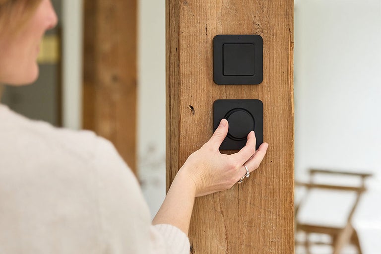 Een persoon bedient een ronde lichtschakelaar of dimmer op een houten balk, waarboven een vierkante lichtschakelaar is gemonteerd.